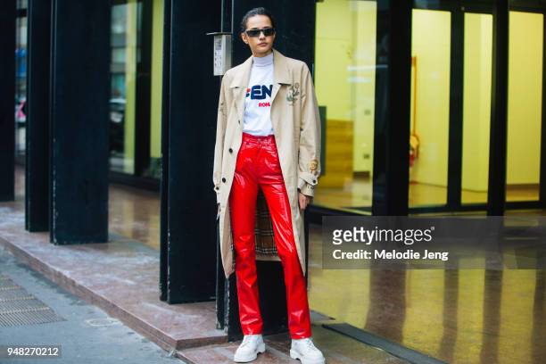 Model Fernanda Oliveira wears a tan trenchcoat with illustrations, a Fendi Roma white roll neck, bright red leather pants, white boots, and...
