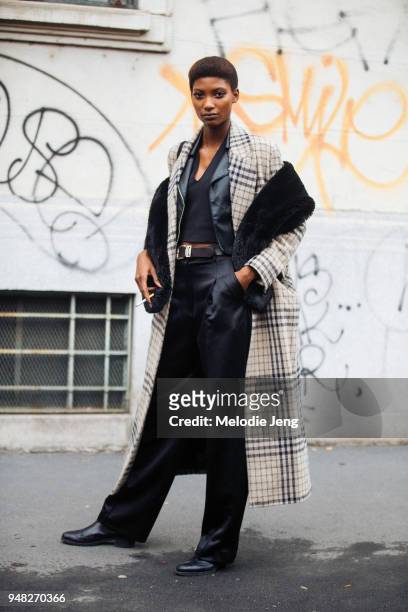 Model Mame Thiane Camara wears a black fur shawl, a tan plaid coat, a black leather jacket, black top, Replay leather belt, black pants, and black...