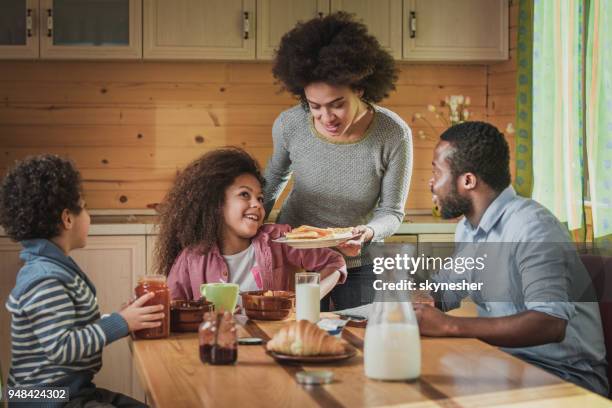 happy black mother serving food to her family in dining room. - african dining stock pictures, royalty-free photos & images