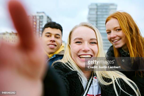 three teenagers taking a selfie on their mobile phone taken from the phone's perspective with hand in view of the camera. two friends lean in from both sides smiling at camera - selfie group stockfoto's en -beelden