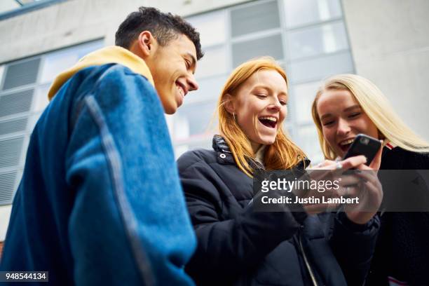 three teens laughing and joking while looking at their phone - young adult fotografías e imágenes de stock
