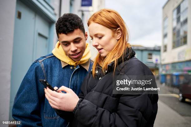teenage couple looking at smartphone and smiling, looking for directions or social media while in the street - couple smartphone stockfoto's en -beelden