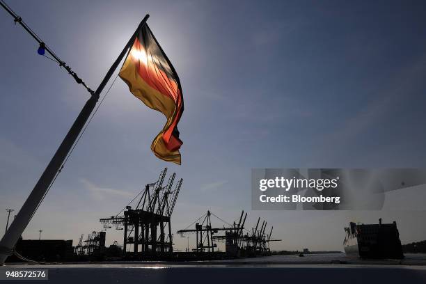 German national flag flies from a sightseeing boat as shipping cranes stand beyond at Terminal Burchardkai in the Port of Hamburg in Hamburg,...
