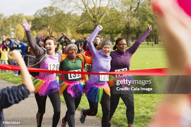 enthusiastic female runners in tutus crossing charity run finish line in park, celebrating - charity benefit stock pictures, royalty-free photos & images