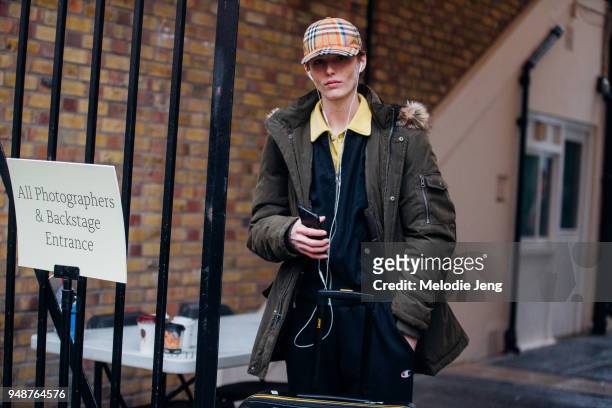 Model Emily Gafford listens to music with headphones and wears a Burberry tartan cap, a green coat, a yellow polo shirt under a blue jacket, and...