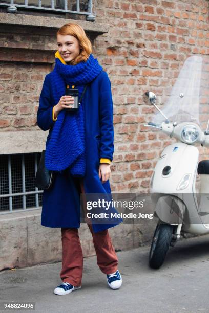 Model Yeva Podurian holds a thermos hot drink and wears a blue coat with an oversized blue knit scarf, maroon pants, and blue sneakers during Milan...