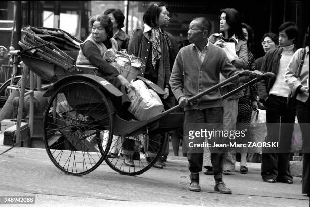 Homme asiatique tirant un pousse-pousse avec une femme dedans et ses sacs de course, 1978 a Hong Kong.