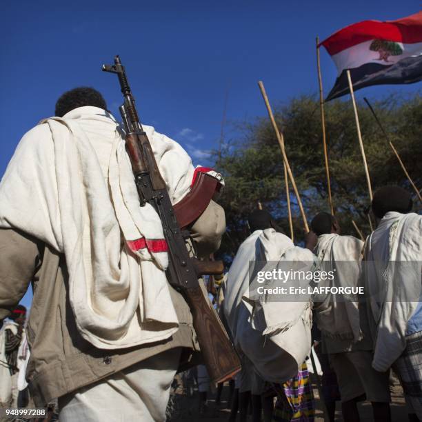 Rear view of a karrayyu tribe man carrying a kalashnikov passing by a group of karrayyu people with oromo flag people during gadaaa ceremony,...