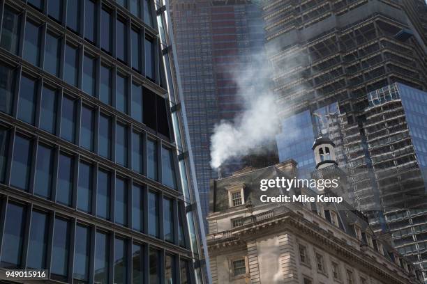 Pollution from the chimney of an old building pours out into the atmosphere amongst modern glass buildings in the City of London, London, England,...