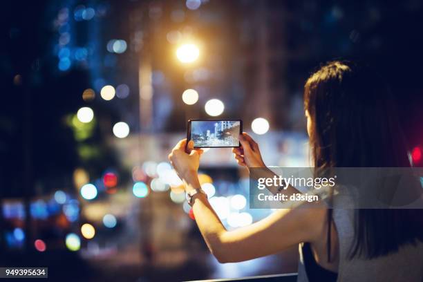 woman capturing city night scene with smartphone against illuminated street lights - woman capturing city night foto e immagini stock