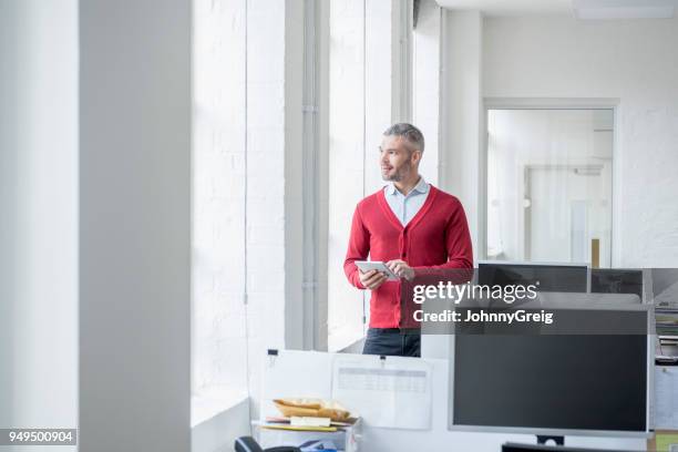 mid adult man in red cardigan using tablet and looking out of window - red cardigan sweater stock pictures, royalty-free photos & images