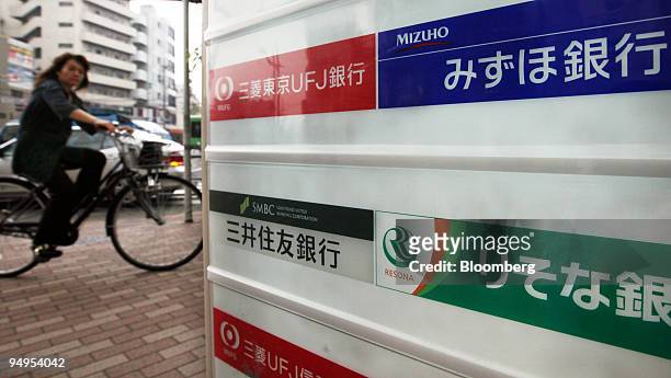 Woman cycles past a board displaying logos for Bank of Tokyo Mitsubishi UFJ, Mizuho Bank Ltd., Sumitomo Mitsui Banking Corp. And Resona Bank Ltd....