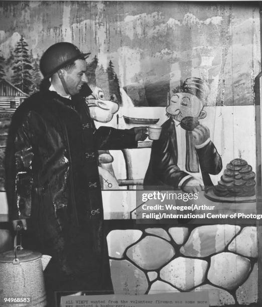 Volunteer fireman, wearing a wet rain coat and helmet, holds a cup to a mural of Wimpy, who is eating a stack of hamburgers, as Popeye looks on, New...