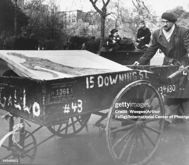 An artist, wearing a beret and trench coat, pushes a pushcart full of paintings, perhaps in Washington Square Park, in Greenwich Village, ca.1942.