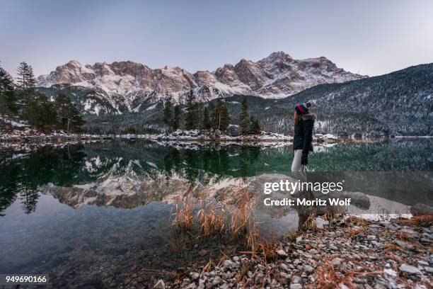 young woman standing on the shore, eibsee lake in winter with zugspitze, spiegelung, wetterstein range, upper bavaria, bavaria, germany - spiegelung foto e immagini stock