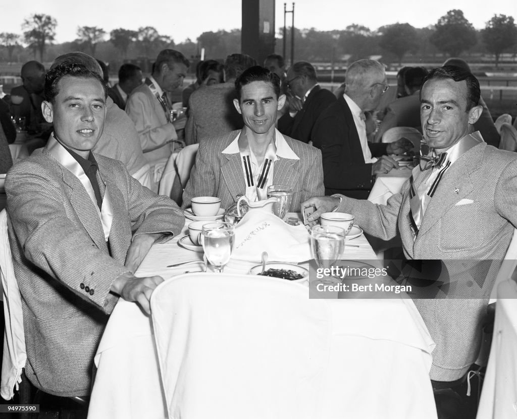 A portrait of jockeys David Gorman, Eric Guerin and Ted Atkinson seated at a table with the racetrack in the background, wearing medals around their necks, Saratoga, New York, c1952