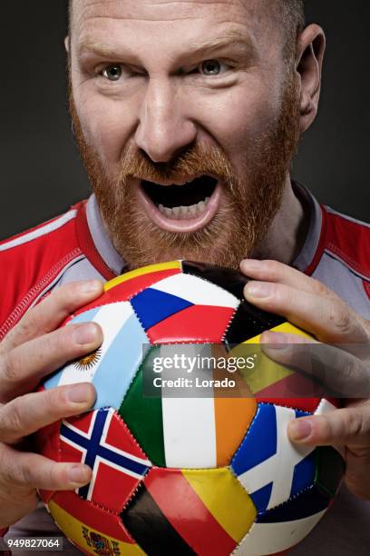 bearded aggressive redhead adult man football player from ireland holding a soccer ball covered in international flags - world cup netherlands stock pictures, royalty-free photos & images