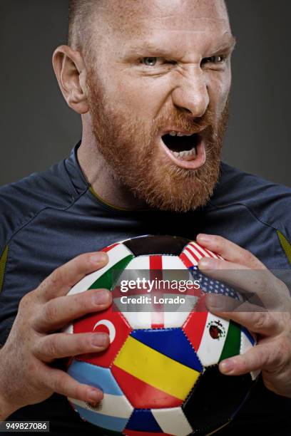 bearded aggressive redhead adult man football player from england holding a soccer ball covered in international flags - world cup netherlands stock pictures, royalty-free photos & images