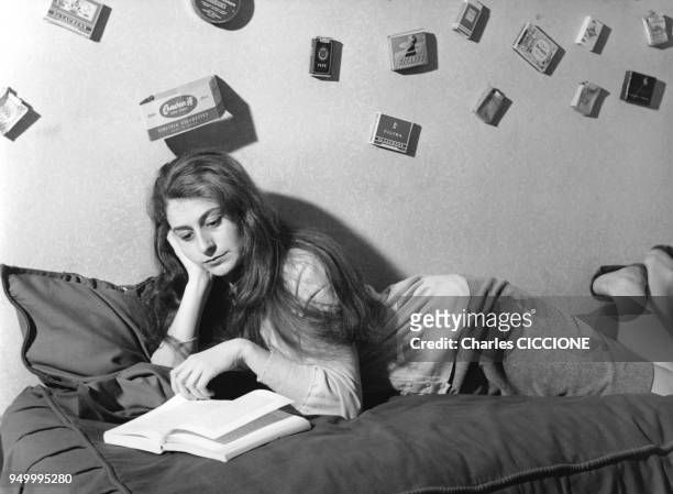 Jeune femme regarde un livre alongee sur son lit, des pochettes d'allumettes decorent le mur derriere elle, circa 1960 a Paris, France.