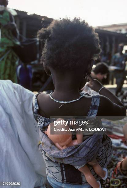 Ue petite fille porte sa poupée dans son dos sur un marché en décembre 1980 en Côte d'Ivoire.