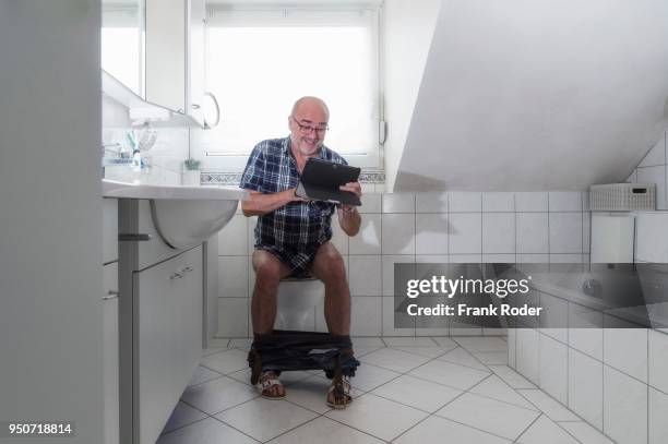 senior man sitting on the toilet, laughing whilst surfing the internet on a tablet, germany - human toilet stock-fotos und bilder