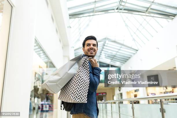 young man holding paper bags and walking in mall - shopping bag imagens e fotografias de stock