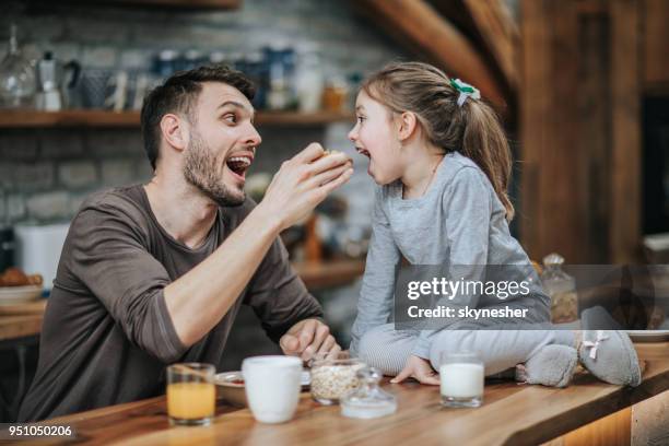 playful father having fun while feeding his small girl in the kitchen. - marmalade sandwich stock pictures, royalty-free photos & images