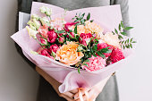 Very nice young woman holding a colourful fresh blossoming flower bouquet of different sorts of roses, carnations, eustoma, peonies on the grey wall background