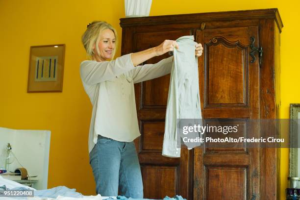 mature woman standing in bedroom, holding out shirt - opbergen stockfoto's en -beelden