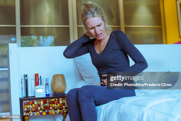 mature woman sitting on bed with hand on stomach and pained expression on face - apendicitis fotografías e imágenes de stock