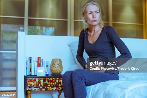 mature woman sitting on bed with hands on stomach and pained expression on face - stomachache fotografías e imágenes de stock