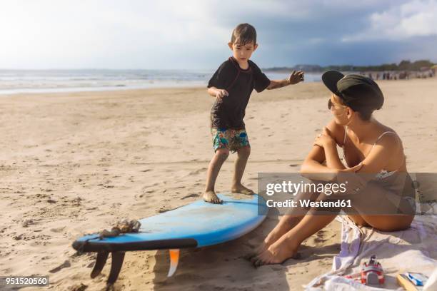 indonesia, bali, boy standing on surfboard, mother sitting on beach - passenger train stockfoto's en -beelden