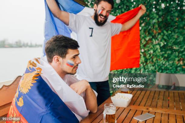 happy french national soccer team supporter standing and holding national flag, his friends supporting russian team sitting down - france national soccer team stock pictures, royalty-free photos & images