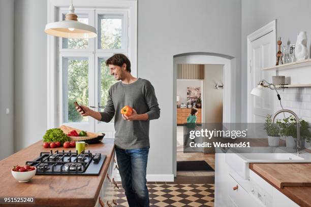 smiling man using smartphone and holding bell pepper in kitchen - bell boy stock-fotos und bilder