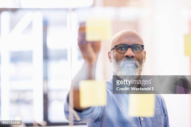portrait of businessman taking adhesive note from glass wall in office - geschäftsstrategie stock-fotos und bilder