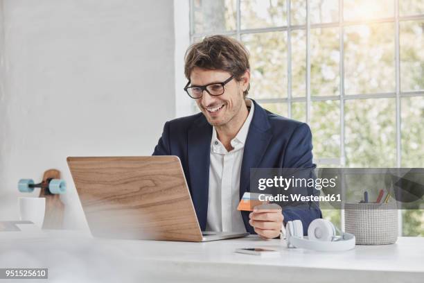 smiling businessman using laptop on desk holding card - business man sitting banking imagens e fotografias de stock