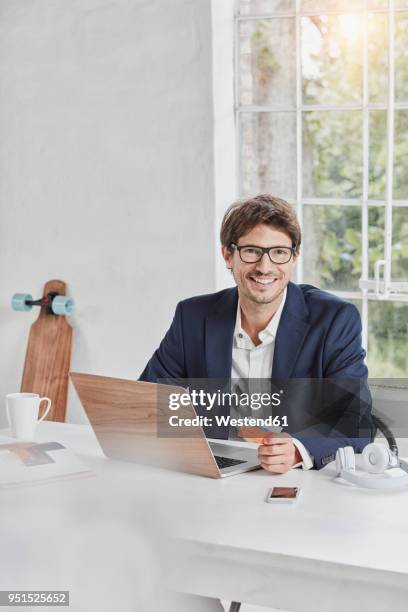 portrait of smiling businessman with laptop on desk holding card - skateboard shop stock pictures, royalty-free photos & images