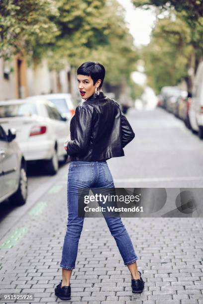 fashionable young woman wearing jeans and leather jacket standing on street - woman lipstick rearview stock-fotos und bilder