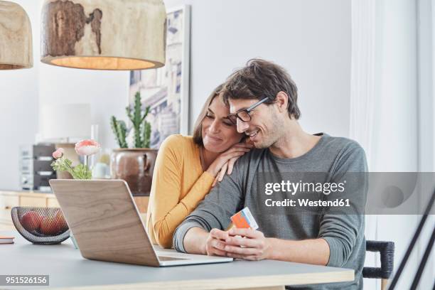 smiling couple with a card using laptop on table at home - mann mit kreditkarte stock-fotos und bilder
