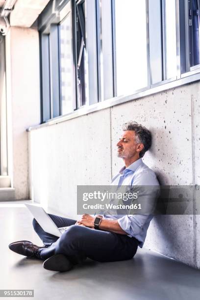mature businessman with laptop sitting on the floor leaning against the wall - schneidersitz stock-fotos und bilder