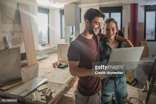 happy embraced couple using laptop while being on construction site in their apartment. - young couple moving house stock pictures, royalty-free photos & images