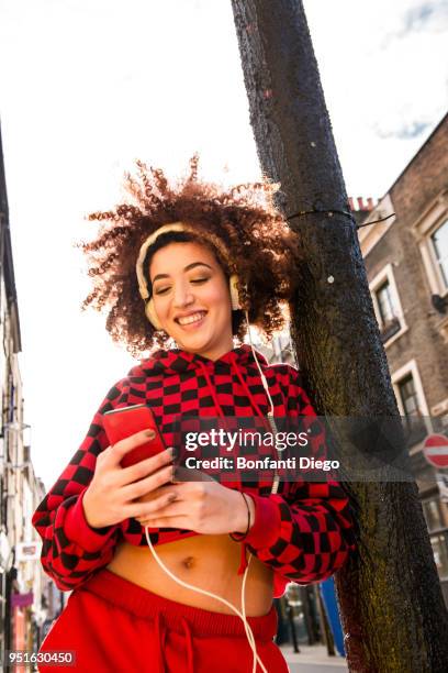 portrait of young woman outdoors, wearing headphones, holding smartphone - leaning against lamppost stock pictures, royalty-free photos & images