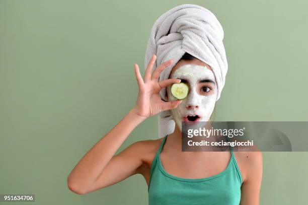 teenage girl with a face mask on holding a cucumber slice in front of one eye - cucumber imagens e fotografias de stock