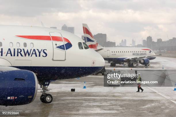 Marshaller wearing yellow high viz tabard and ear-defenders standing with British Airways BAW Airbus A318-100s parked in the snow.