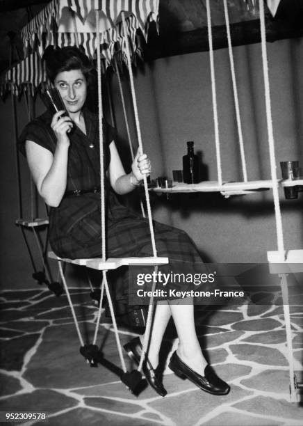 Jeune femme assise sur un siège suspendu où les bouteilles ainsi que le comptoir sont également suspendus, mobilier appelé 'bar-balançoire' présenté...