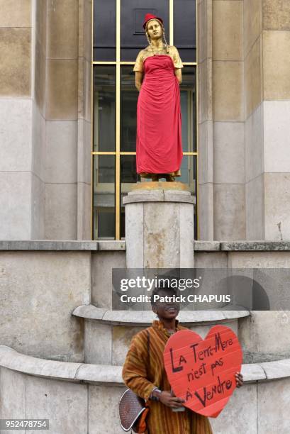 Femme tenant une pancarte en forme de coeur 'La terre mère n'est pas à vendre' devant une statue habillée avec une robe et une coiffe rouge lors de...