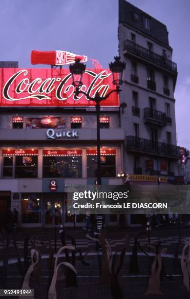 Place Blanche in Montmartre with a huge neon sign for Coca-Cola on one of the buildings and underneath, a Quick fast food store. In the foreground is...