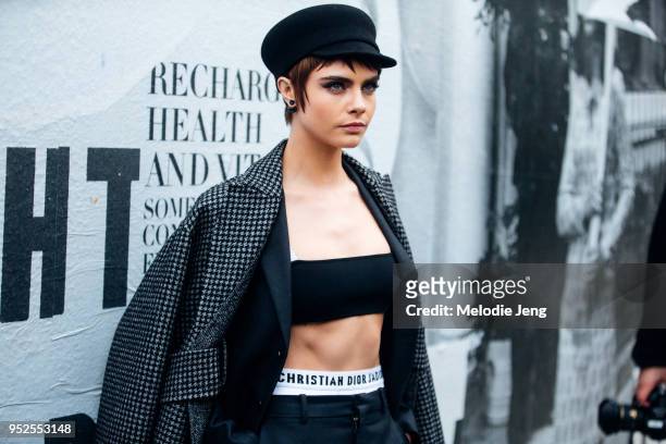 Model Cara Delevingne wears a black conductor hat, gray houndstooth coat, black blazer, Christian Dior bra top and underwear, and black trousers at...