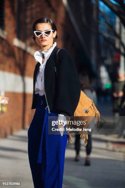Model Dipti Sharma wears futuristic-style triangular white sunglasses, a black top, brown purse with a fringe, and high-waisted blue trousers on...