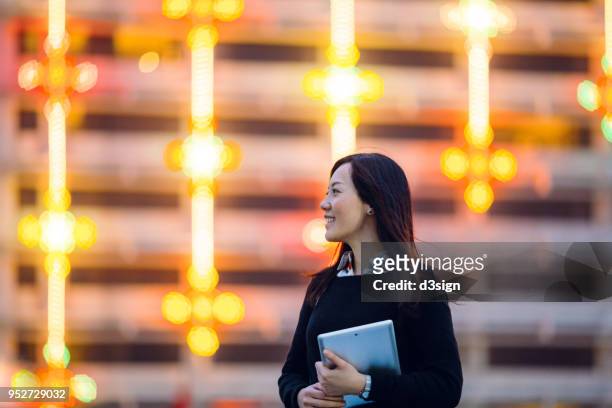 portrait of young woman holding digital tablet outdoors against illuminated highrise building - businesswear stock pictures, royalty-free photos & images